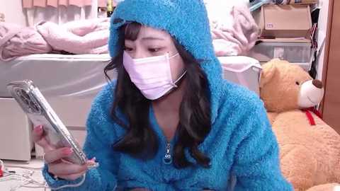 Media: Video of an Asian woman with long black hair, wearing a blue fuzzy hooded bathrobe and a white medical mask, seated on a floor, reading a tablet. Background includes a bed with pink bedding, a teddy bear, and a shelf.