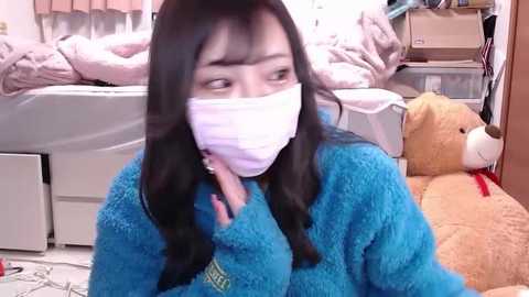 Media: Video of an Asian woman with long black hair, wearing a blue fleece jacket, white mask, and a beige teddy bear, in a cluttered room with a bed, boxes, and a laundry basket.