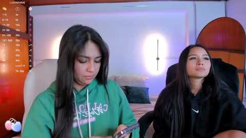 Media: A video of two young women with long black hair, one in a green hoodie, the other in black, sitting on a bed in a modern, brightly lit bedroom.