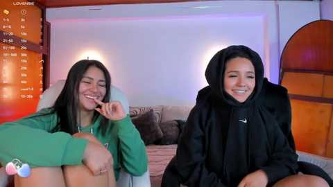 Media: Video of two women with long black hair, one in a green hoodie and the other in a black hijab, sitting on a couch in a cozy, dimly-lit room, smiling.