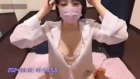 Media: Video of an Asian woman with light skin, wearing a white lab coat unbuttoned to reveal a pink lace bra, seated in a chair, adjusting her mask, in a clinical setting.