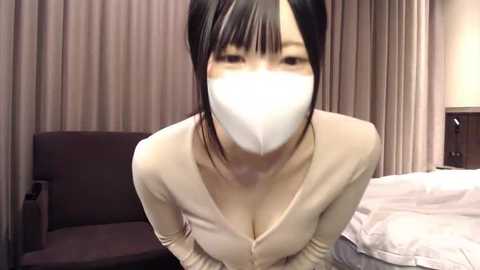 Media: Video of an East Asian woman with long black hair, wearing a white face mask, beige top, and sitting on a bed with white sheets, in a dimly-lit bedroom with beige curtains.