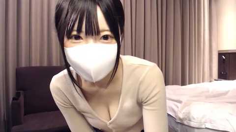 Media: A video of an Asian woman with straight black hair, wearing a white surgical mask and beige long-sleeved top, leaning forward in a dimly-lit room with beige curtains and a white bed.