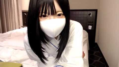 Media: Video of an Asian woman with long black hair, wearing a white face mask, white lab coat, and a stethoscope, leaning over a hospital bed in a dimly lit room with beige walls and a dark carpet.