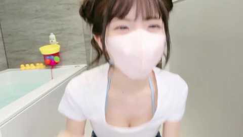 Media: A blurred video of a young Asian woman with fair skin, wearing a white face mask, white top, and blue jeans, standing in a modern bathroom with a bathtub, colorful bath toys, and grey tiled walls.