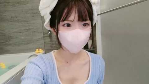 Media: Video of a young Asian woman with straight black hair and bangs, wearing a light blue sweater, white face mask, and white headband, standing in a modern bathroom with gray tiled walls and a bathtub filled with yellow bath bombs.