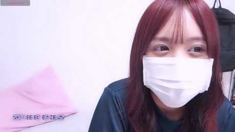 Media: Video of a woman with long, reddish-brown hair, wearing a white medical mask, dark blue shirt, and pink towel, in a sterile, white-walled medical setting.