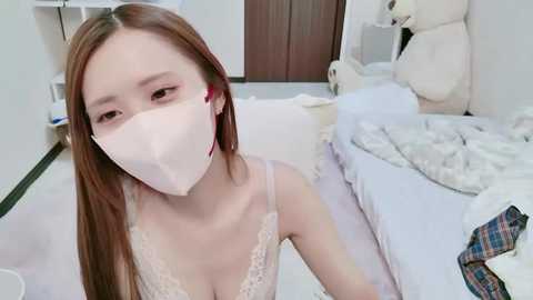 Media: A video of a young Asian woman with long, straight brown hair, wearing a white mask, lace bra, and sitting on a bed in a minimalist, white bedroom with plush toys and a plaid shirt.