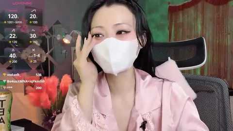 Media: Video of an Asian woman with long black hair, wearing a pink satin robe and white surgical mask, seated in a dark chair with a blurred background of a room with a green wall and a wooden curtain.