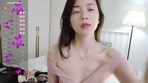 Media: Video of an Asian woman with straight black hair, wearing a pink off-shoulder top, in a modern, white room with a lamp and bed.