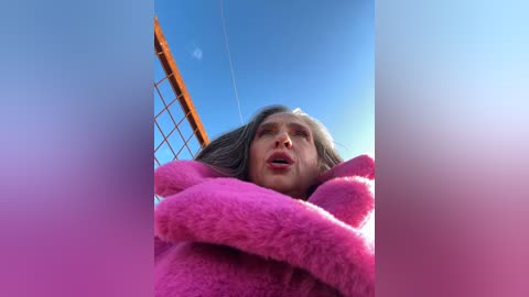 Media: A video of a woman with long gray hair, wearing a bright pink fur coat, standing in front of a blue sky with a yellow-orange tower in the background.