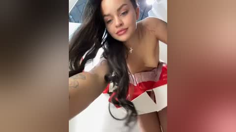 Media: Video of a young woman with long black hair, light skin, and red lipstick, wearing a strapless red dress with white accents, taking a selfie, surrounded by blurred faces.