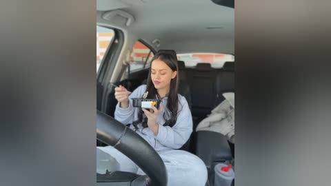 Media: Video of a young woman with long black hair, wearing a light grey hoodie, eating a bowl of food in a car. The car's interior is clean and modern.