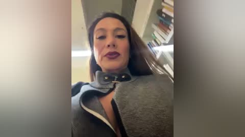 Media: Video of a middle-aged woman with fair skin, dark hair, and bold makeup, wearing a gray trench coat with a black belt, standing indoors near a bookshelf.