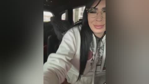 Media: A video of a woman with long black hair, wearing glasses and a white hoodie with a graphic design, taking a selfie from the backseat of a car. The background shows dark car upholstery and blurred cityscape.