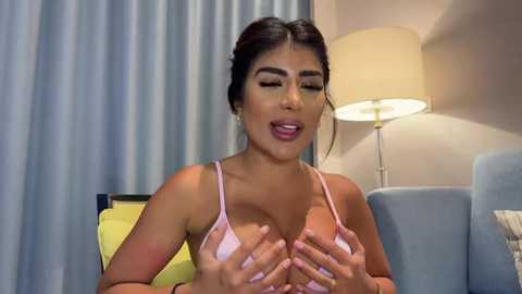 Media: Video of a Latina woman with medium-dark skin, dark hair, and full lips, wearing a pink bra, covering her breasts with her hands, in a modern hotel room with blue curtains and a beige lamp.