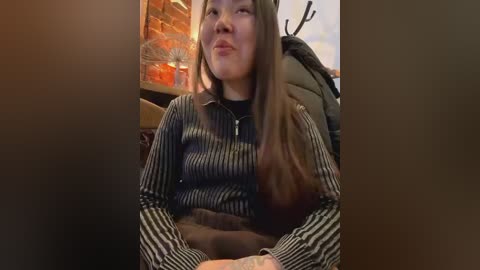 Media: Video of an Asian woman with long brown hair, wearing a striped sweater, sitting on a couch in a warmly lit room with brick walls and a deer head on the wall.