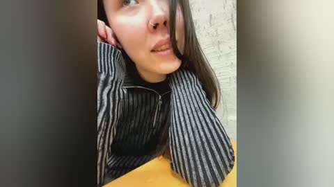 Media: Video of a young woman with fair skin, long brown hair, and a nose ring, wearing a black and white striped jacket, leaning against a brick wall.