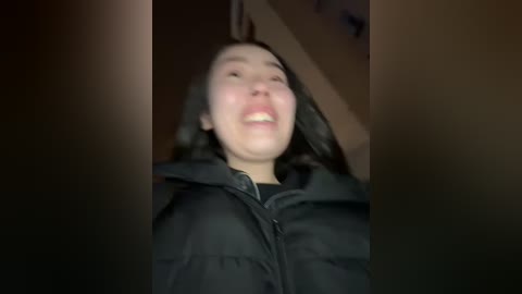 Media: A blurry video of a smiling Asian woman with dark hair and fair skin, wearing a black puffer jacket, taken indoors.