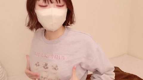 Media: Video of a woman with short, dark brown hair, wearing a light purple \"Witcher\" t-shirt, white face mask, and brown pants, seated on a white bed.