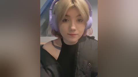 Media: A video of a young woman with pale skin and blonde hair, wearing large purple headphones, a black turtleneck, and a gray jacket, with blurred background.