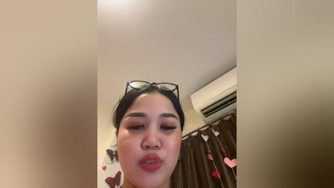 Media: A video of a young Asian woman with medium skin tone, wearing black-rimmed glasses and red lipstick, blowing a kiss. She stands in a room with a white ceiling, brown curtains adorned with pink hearts, and an air conditioning unit.