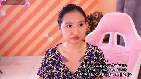 Media: A video of a young Asian woman with dark hair tied back, wearing a black and white patterned blouse. She sits in a pink gaming chair in a room with orange and white striped wallpaper. Korean text is overlaid.