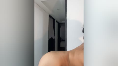 Media: A video of a nude, light-skinned woman's backside, partially obscured by a white wall. The background shows a modern, minimalist bedroom with a white ceiling and dark furniture.