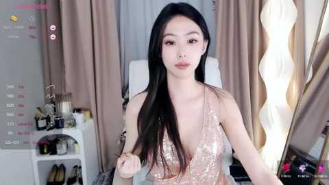 Media: Video of a young Asian woman with long black hair, wearing a low-cut, glittery gold dress, standing in a modern, well-lit room with beige curtains and a white chair.