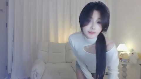 Media: A young woman with long black hair, fair skin, and a slender build, wearing a white off-shoulder top, sits on a white couch in a softly lit room with white curtains and a table lamp.