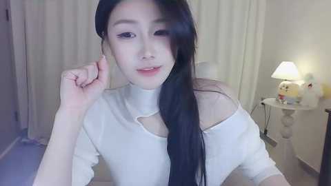 Media: Video of an East Asian woman with long black hair, fair skin, and wearing a white off-shoulder top, smiling, in a softly lit room with a table lamp and white curtains.