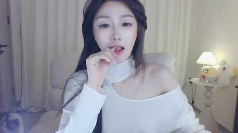 Media: A video of an East Asian woman with long black hair, wearing an off-shoulder white sweater, biting her finger in a bedroom with white curtains and a lamp.