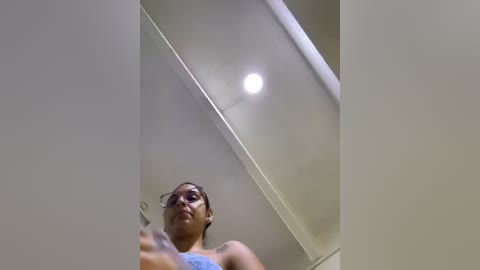 Media: Video of a young woman with dark skin, wearing glasses and a blue tank top, taking a selfie from a low angle, capturing the ceiling light in the background.