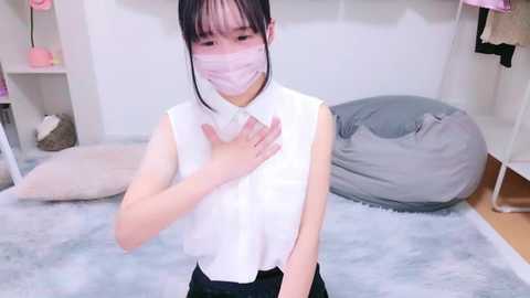 Media: A video of an East Asian woman with fair skin, straight black hair, and a white mask, wearing a sleeveless white blouse, standing in a light-colored room with gray and white cushions.