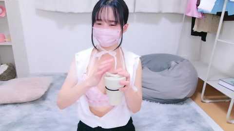 Media: Video of an Asian woman with pale skin and dark hair wearing a white blouse, pink face mask, and gloves, applying lotion to her face in a cozy, cluttered room with a gray beanbag, shelves, and clothes.