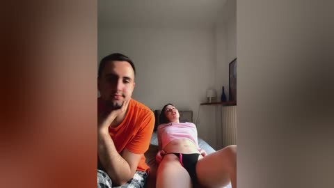 Media: Video of a young couple in a bedroom, man in orange t-shirt, woman in pink top, lying on bed, soft lighting, simple decor.