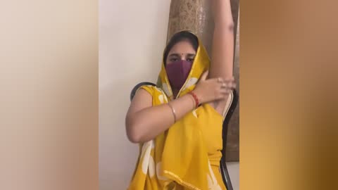 Media: A video shows a South Asian woman in a yellow sari, partially covering her face with a maroon mask, stretching her right arm upwards, set against a beige wall and textured brown curtain.