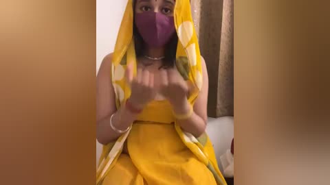 Media: Video of a South Asian woman with medium brown skin, wearing a yellow sari, purple mask, and silver jewelry, gesturing with her hands.