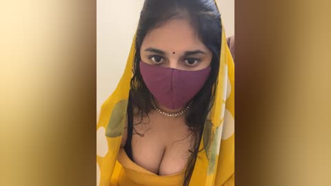 Media: Video of a South Asian woman with medium skin tone, wearing a yellow saree with white polka dots, a purple face mask, and a gold necklace. She has long, dark hair and is looking directly at the camera.