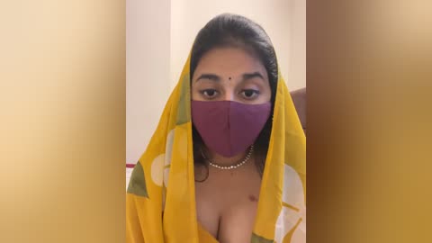 Media: Video of a South Asian woman with medium-dark skin, wearing a yellow sari with a purple face mask, a pearl necklace, and a bindi. She has long, dark hair and is indoors against a beige background.