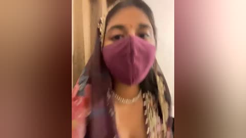 Media: A video shows a woman in a maroon mask, floral headscarf, and traditional attire, indoors with blurred background.