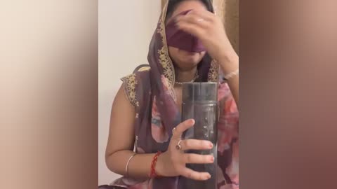 Media: Video of a South Asian woman in a maroon sari, covering her face with one hand, holding a glass of water.