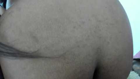 Media: A close-up video of a person's lower back and buttocks, showing light brown skin, a few moles, and a small, faint scar. The background is out of focus with a light-colored wall and a soft, textured surface.