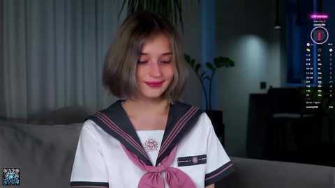 Media: A video of a young girl with short, light brown hair, wearing a school uniform with a pink bow, smiling softly. She sits in a dimly lit living room with a gray sofa and a plant in the background.