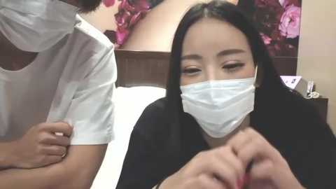 Media: Video of an Asian woman with long black hair and a surgical mask, wearing a black top, looking down as a person with short hair in a white shirt adjusts her mask. Background features a floral wallpaper and a wooden headboard.