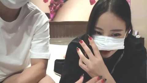 Media: Video: Asian woman with long black hair and red nails in surgical mask, sitting beside a man in a white shirt, both indoors, blurred background.