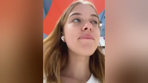 Media: Video of a young Caucasian woman with light skin, straight brown hair, and blue eyes, wearing a white top, looking up thoughtfully. Background features a red and blue patterned object.