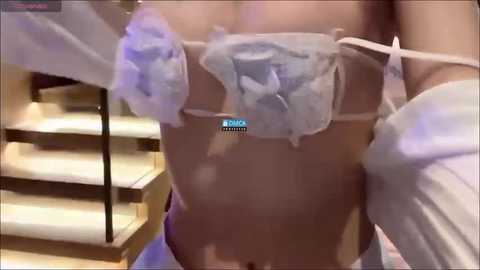 Media: A video of a woman wearing a sheer, strapless, light blue bra adorned with large, fluffy rose appliqu\u00e9s. Her midsection is partially visible, revealing a toned, lightly tanned abdomen. Background includes beige carpeted stairs.