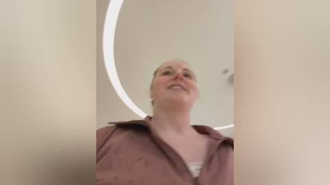 Media: A video of a smiling woman with fair skin and short blonde hair, wearing a pink jacket, standing in a modern, circular room with a bright, circular light fixture in the background.