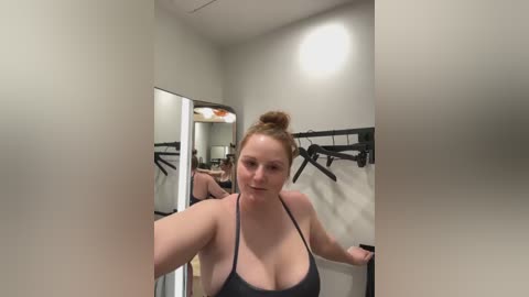 Media: Video of a fair-skinned, red-haired woman with a bun, wearing a black halter top, standing in a gym locker room with mirrors, black hooks, and white walls.